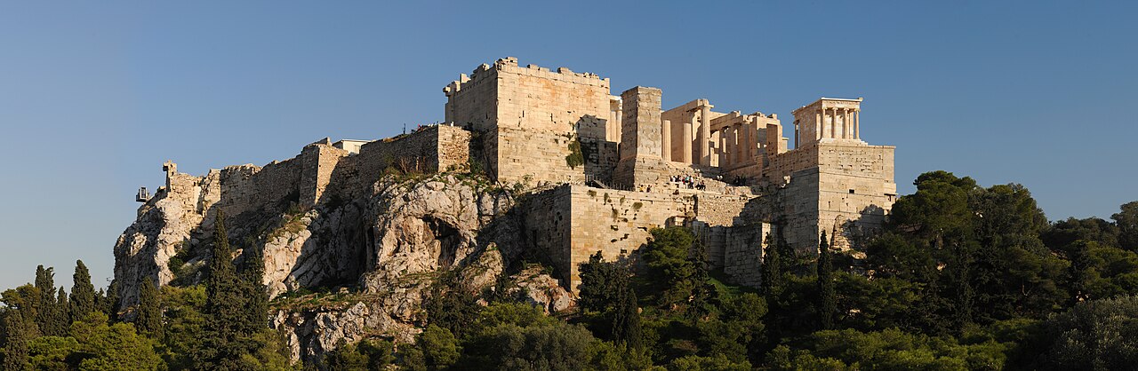 Sunlit stone walls and monuments of the Acropolis rising above trees, seen from the Areopagus in Athens.